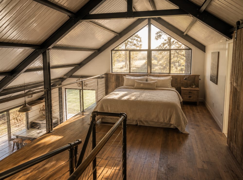 Cama en altillo enmarcada por cerchas de acero y luz de ventana de hastial